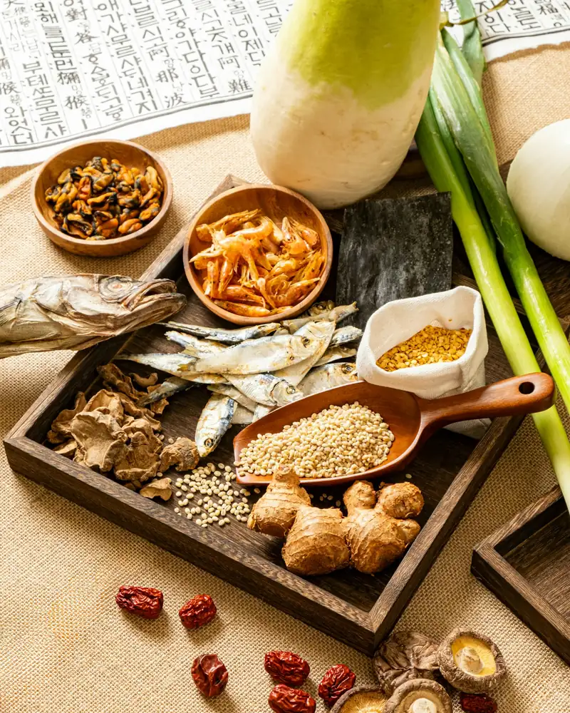 Traditional Korean medicinal ingredients on a wooden tray: jujube, shiitake, ginger, barley, and dried anchovy, on a classical Korean text
